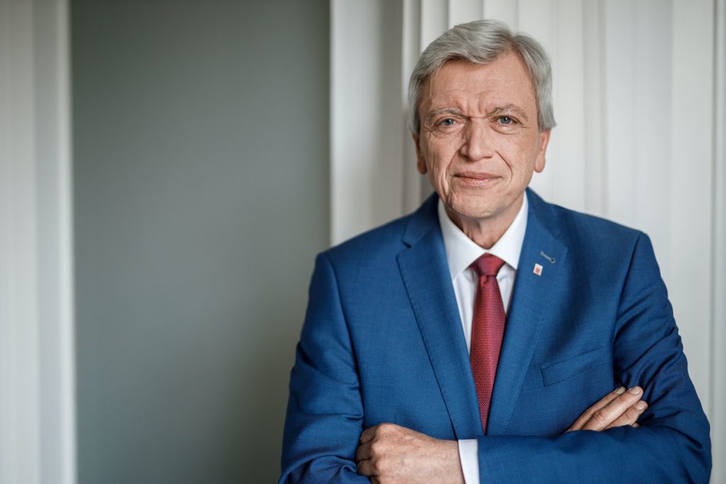 Volker Bouffier steht in blauem Anzug mit weißem Hemd und roter Krawatte lächelnd vor einer grauen Wand und blickt in die Kamera.