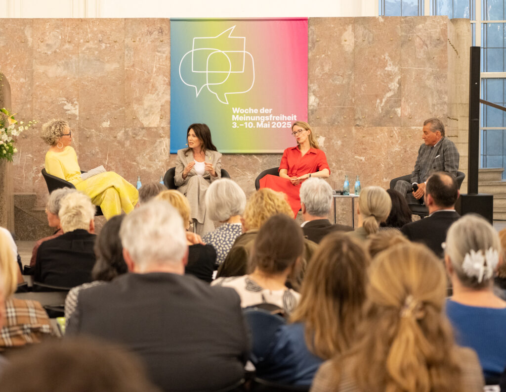 Podiumsdiskussion in hellem Saal: Vier Personen sitzen vor Publikum; mittig farbiges Banner „Woche der Meinungsfreiheit 3.–10. Mai 2025“; große Blumendeko in vorwiegend Gelbtönen links.