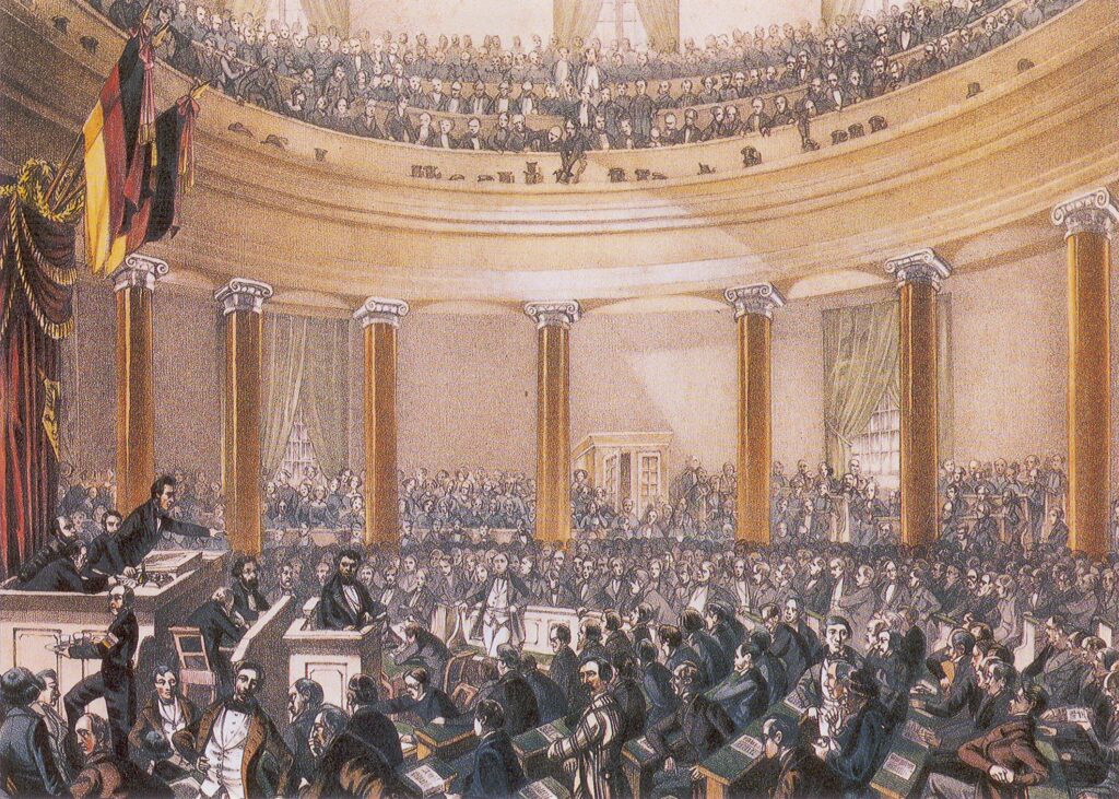 Historische Darstellung der Frankfurter Nationalversammlung 1848 in einer großen Säulenhalle mit zahlreichen versammelten Abgeordneten und Zuschauenden. Links hängen Fahnen in schwarz-rot-gold.